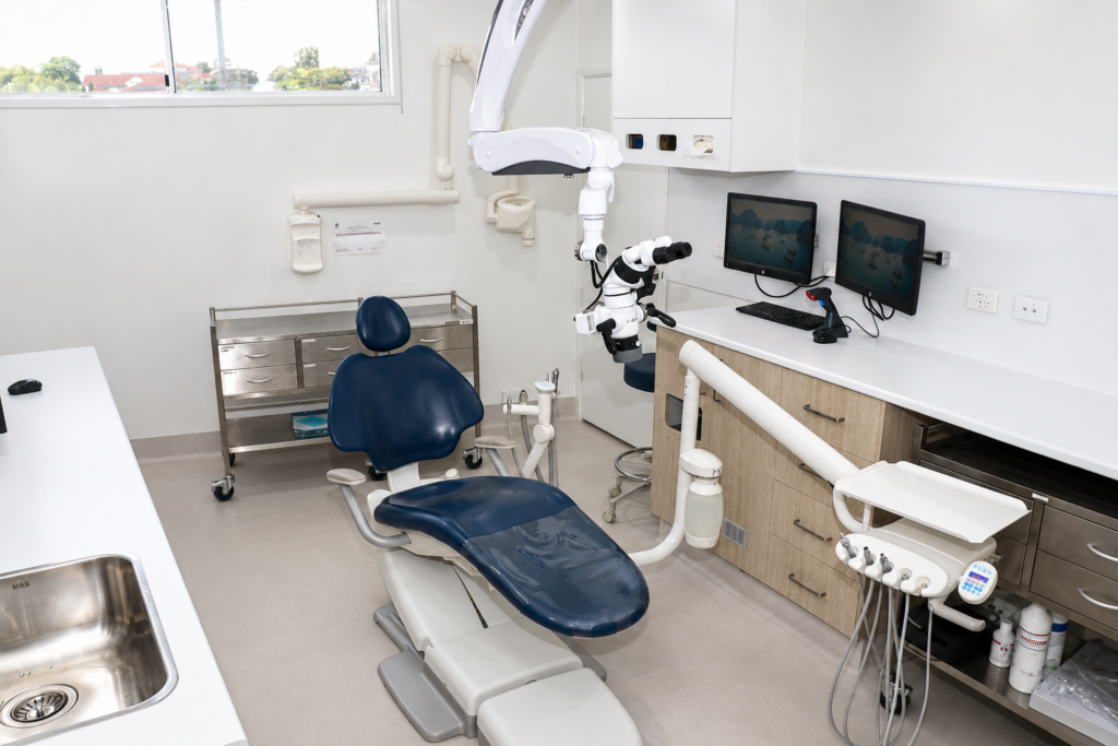 Modern dental operatory with a blue patient chair, overhead robotic microscope, and two monitor screens on the counter.