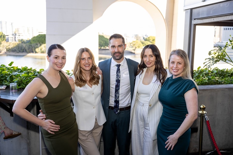 Five professionals in business attire pose together on an outdoor terrace with a river and cityscape in the background, smiling for a group portrait.