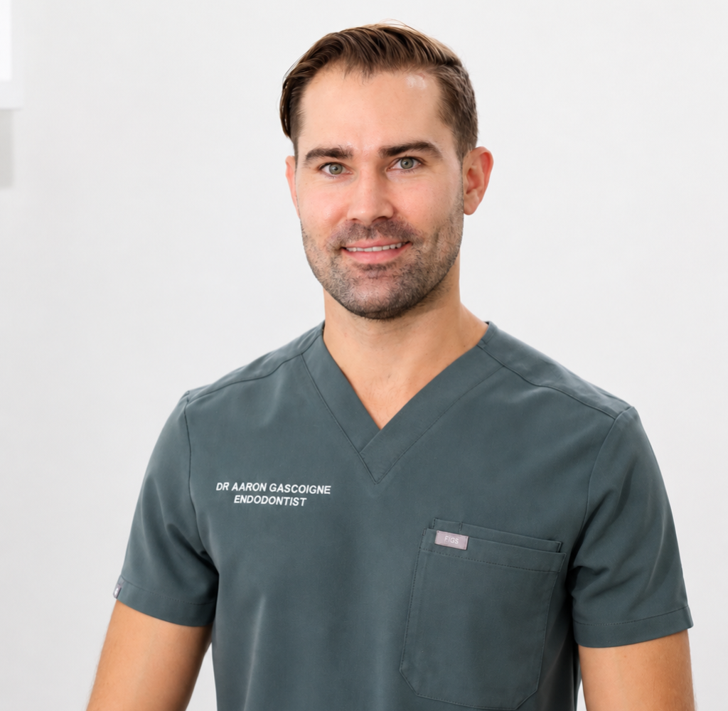 Male dentist in gray scrubs smiling at camera; uniform reads 'Dr Aaron Gascogne, Endodontist' on chest.