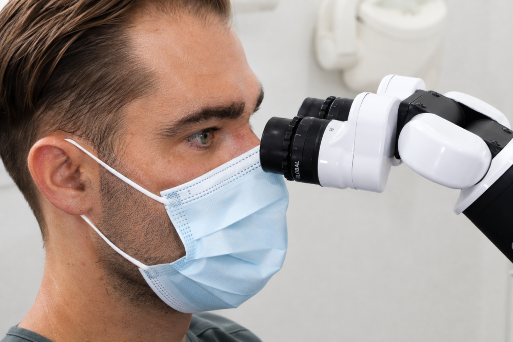 Man wearing a blue surgical mask looks into a white binocular microscope in a lab setting.